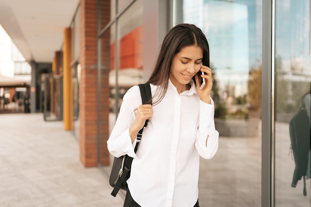 Woman with business backpak walking while speaking on the phone near a a business building