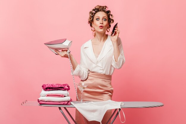 woman with curlers in surprise looks at front, talking on phone and holding iron