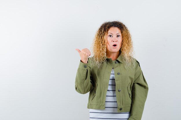 Woman with curly blonde hair in green jacket pointing aside with thumb and looking wondered , front view.