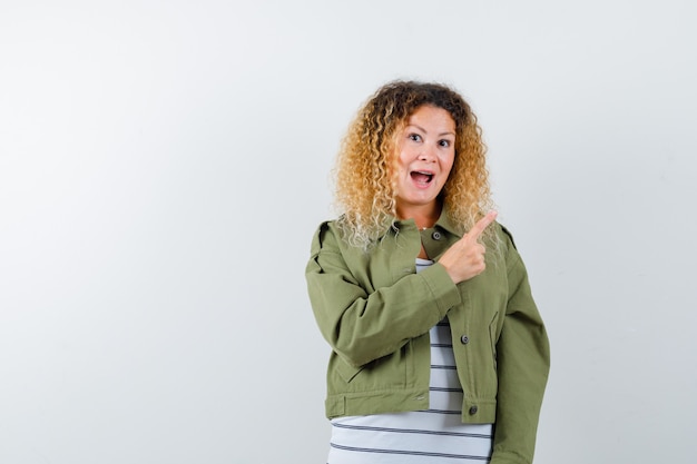 Woman with curly blonde hair pointing at upper right corner in green jacket and looking happy , front view.