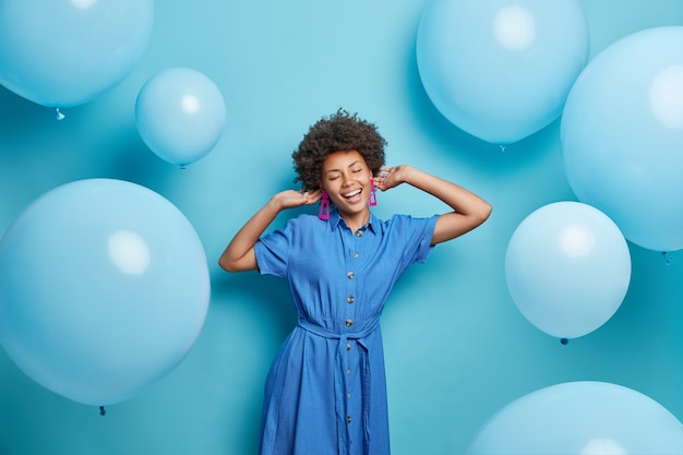 Free Photo woman with curly hair dressed in fashionable dress enjoys music and party poses around inflated balloons has festive moos isolated on blue