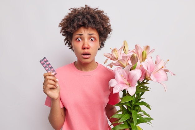 Free photo woman with curly hair holds bouquet of lily flowers has allergic reaction to pollen holds medicaments to cure symptoms of disease wears pink t shirt isolated on white