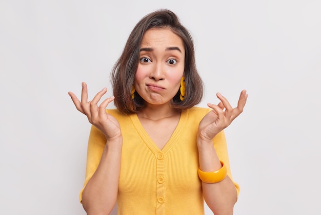 woman with dark hair clueless expression raises hands shrugs shoulders with bewilderment doesnt know how to act wears casual yellow jumper isolated on white