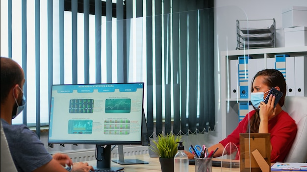 Free photo woman with face mask talking on phone looking at desktop and analyzing the statistics. freelancer working in workplace chatting with remotely team speaking on smartphone in front of computer