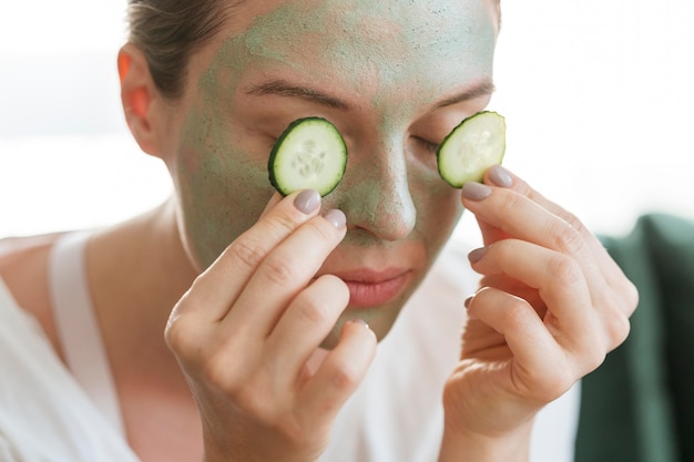 Woman with facial mask putting slices of cucumber