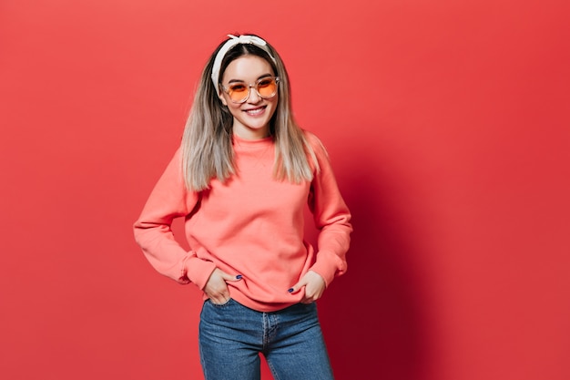 Free Photo woman with hair band, wearing orange glasses and sweatshirt, posing on red wall