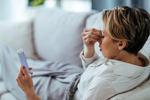 Woman with headache holding her head in pain while relaxing on the sofa and using mobile phone