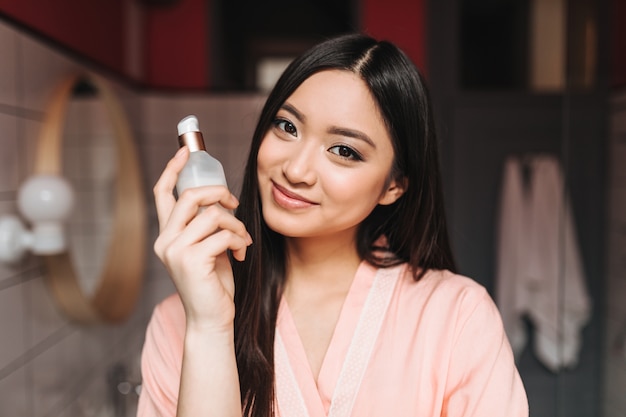 woman with healthy skin is cute smiling and posing with face cream on wall of bathroom