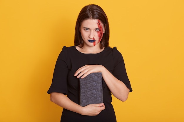 woman with horror Halloween make up and bloody wound posing in studio on yellow, young female with dengerous sight holds book with incantation, dresses black dress