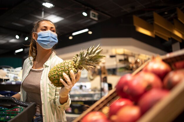 Woman with hygienic mask and rubber gloves and shopping cart in grocery buying fruit during corona virus and preparing for a pandemic quarantine