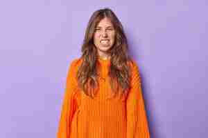 Free photo woman with long dark hair clenches teeth with irritation squints face being annoyed with something wears casual orange knitted sweater isolated on purple