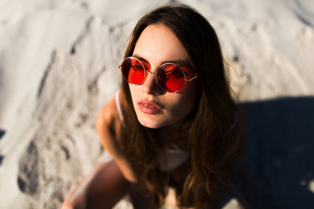 Woman with long hair in red sunglasses sits on white sand