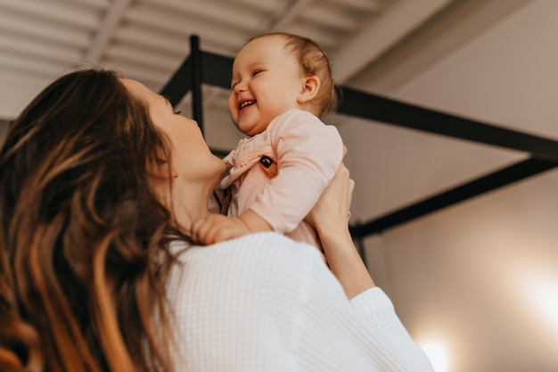 Free photo woman with long hair tickles and throws up laughing baby in light-colored home sweater.