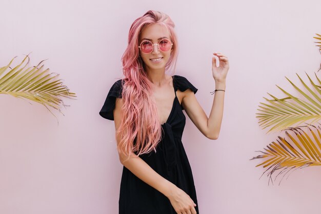 woman with long pink hair standing beside exotic palm trees in studio.