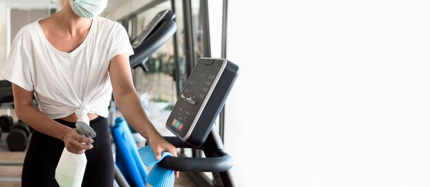 Woman with medical mask cleaning gym equipment with copy space