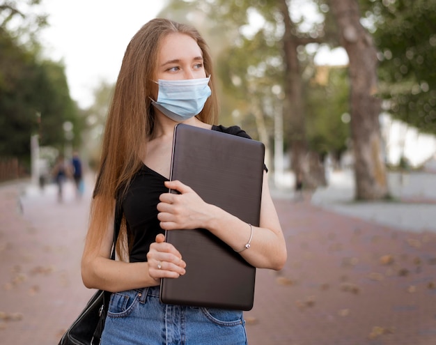 Free Photo woman with medical mask holding a laptop outside