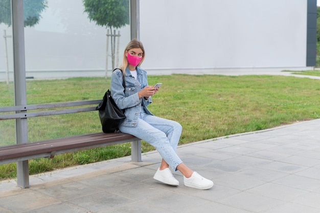 Woman with medical mask using smartphone while waiting for the public bus