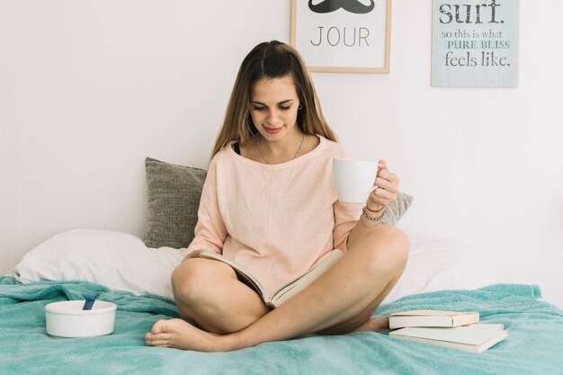 Woman with mug reading book on bed