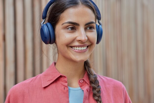 woman with pigtail has white teeth listens audio track in wireless headphones wears pink shirt poses