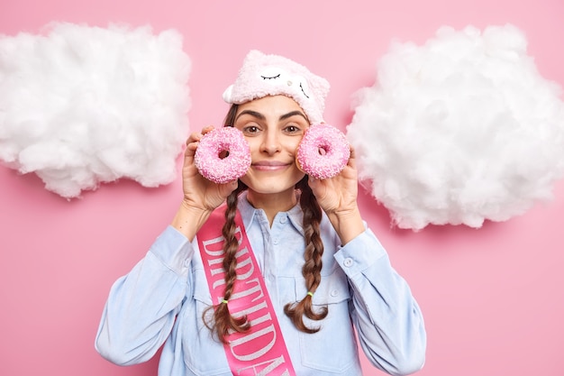 Free photo woman with pigtails holds glazed doughnuts near face wants to eat bakery product wear sleepmask on forehead shirt and ribbon written word birthday poses around white clouds