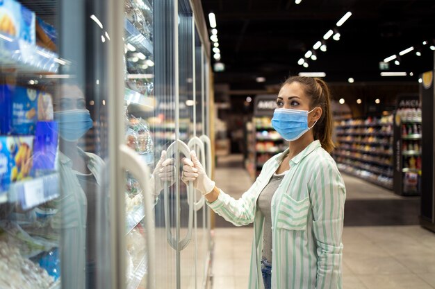 Woman with protective mask and gloves opening freezer in supermarket during COVID-19 pandemic or corona virus