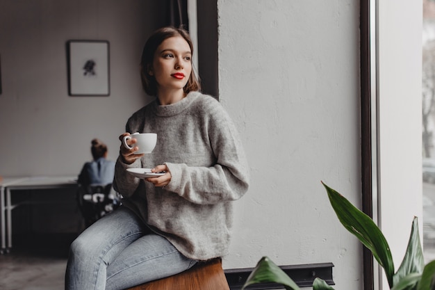 Free photo woman with red lipstick dressed in cashmere sweater is sitting on windowsill with cup of coffee on background of working girl in office.