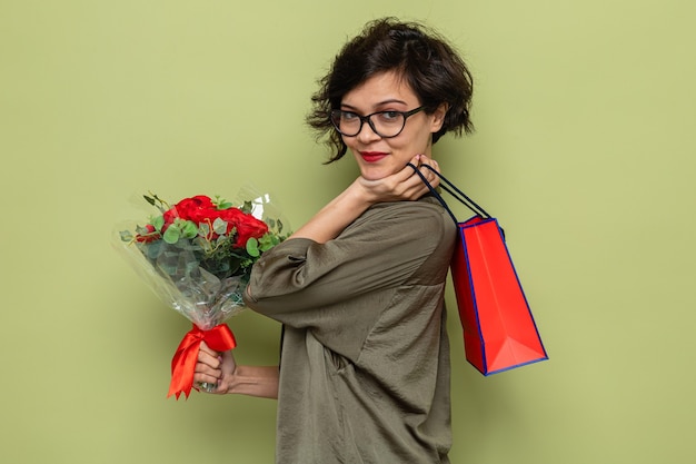 Woman with short hair holding bouquet of flowers and paper bag with gifts looking happy and pleased