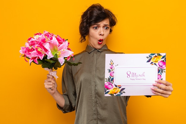 Free photo woman with short hair holding greeting card and bouquet of flowers looking at card confused and surprised celebrating international women's day march 8