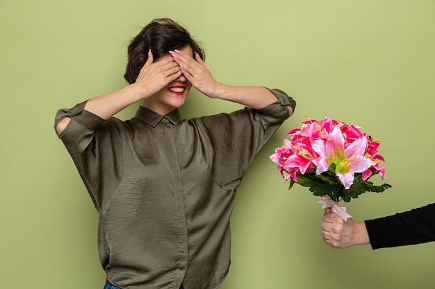 Woman with short hair looking surprised covering eyes with hands while receiving bouquet of flowers from her boyfriend celebrating international women's day march 8 standing over green background