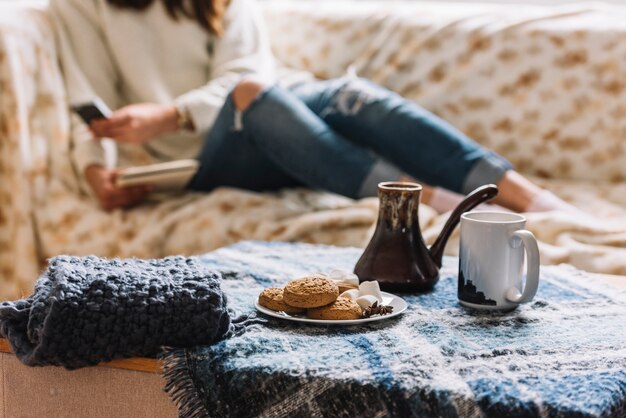 Woman with smartphone on sofa near table with drink and cookies