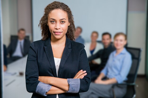 Woman with suit sitting at a table with partners behind and arms crossed