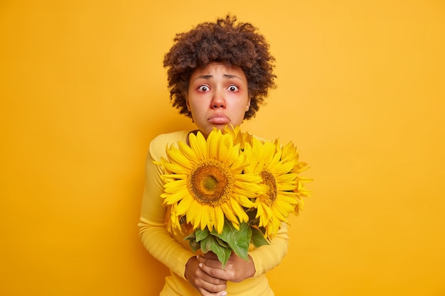 Free photo woman with symptoms of allergy has red swollen eyes holds bouquet of sunflowers isolated on vivid yellow