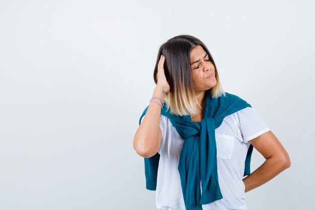 Woman with tied sweater keeping hand on head in white t-shirt and looking pensive. front view.