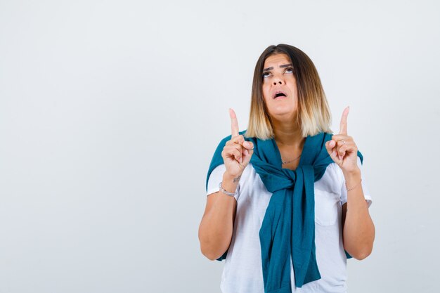 Woman with tied sweater pointing up, looking upward in white t-shirt and looking hopeful , front view.