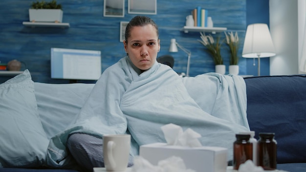 Woman with virus infection sitting in blanket looking at camera. Sick person feeling cold and shivering because of seasonal flu at home. Adult with illness and treatment against disease