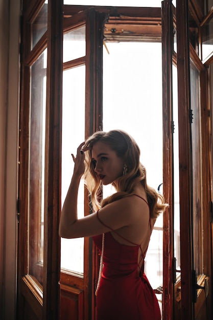 Free photo woman with wavy hair dressed in festive red outfit posing near window curly blond lady in silk dress looking into camera