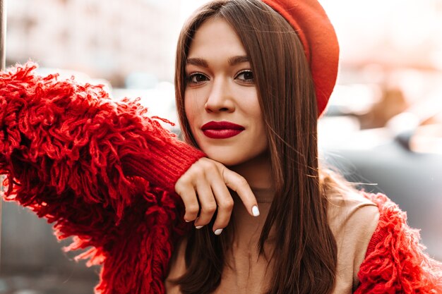 Woman with white manicure and red lips is looking at camera against background of street. woman dressed in red beret and wool jacket leaned on window.