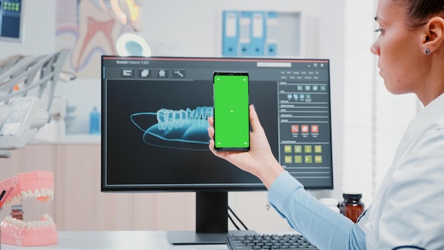 Woman working as dentist with green screen on smartphone in oral care office. Dentist holding mobile phone with mockup template and isolated background for teethcare and dentistry.