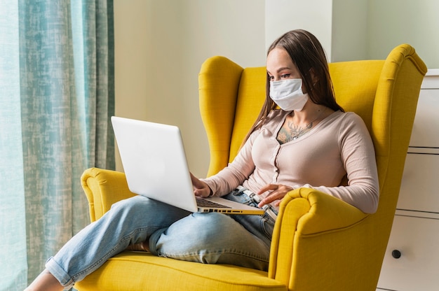 Woman working on laptop from armchair during the pandemic while wearing medical mask