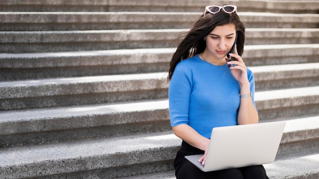 Woman working on laptop outdoors on steps with smartphone