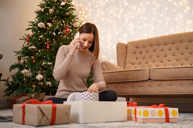 Woman wrapping Christmas gift and talking on the phone while sitting in Christmas