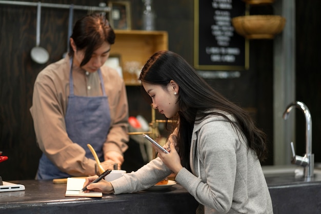 Woman writing on a notebook while man is cooking in a restaurant