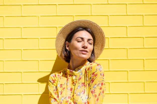 Free photo woman in yellow summer dress and hat on yellow brick wall calm and positive, enjoys sunny summer days