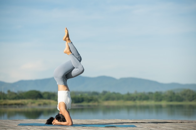 Woman on a yoga mat to relax in the park. Young sporty asian woman practicing yoga, doing headstand exercise, working out, wearing sportswear, pants and top.