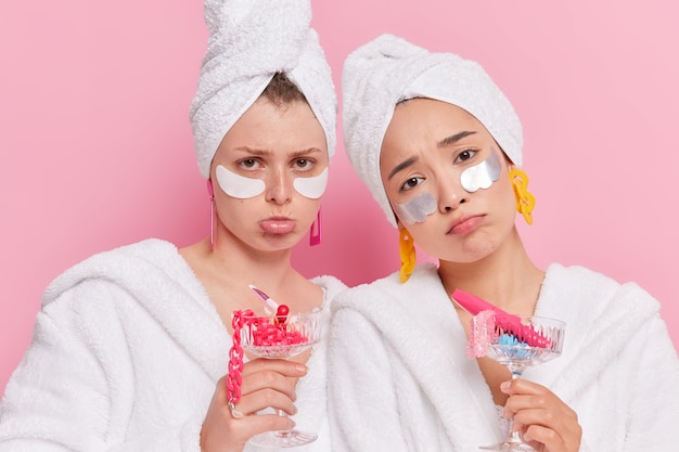 Free photo women apply beauty patches to moisturize skin hold cocktail glasses full of different accessories being disappointed by something wear bathrobes towel on heads isolated on pink wall