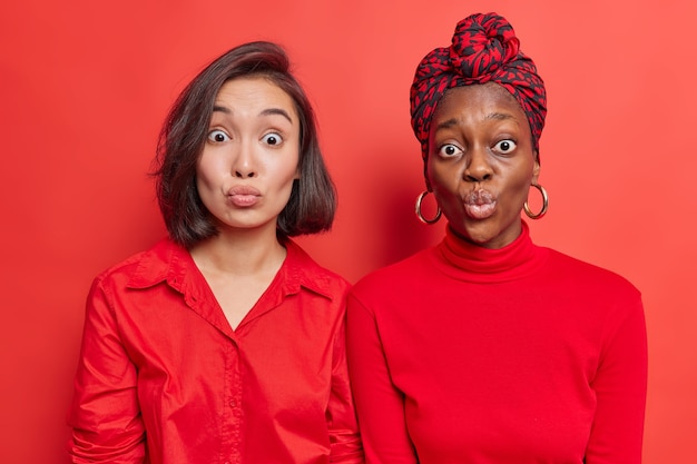 women best friends stand closely to each other keep lips rounded wait for kiss wears red clothes pose on bright studio wall. Mixed race female models with puckered lips. Face expressions