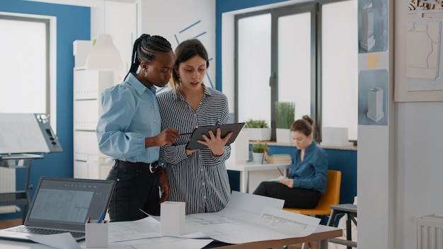 Free photo women looking at digital tablet and blueprints to plan building layout for construction. colleagues working together to design structure and architectural development. architects at work