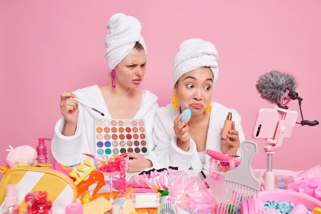 Free photo women record tutorial video apply eye shadow and foundation on face dressed in domestic clothes sit at messy table with cosmetic products give detailed review. social media