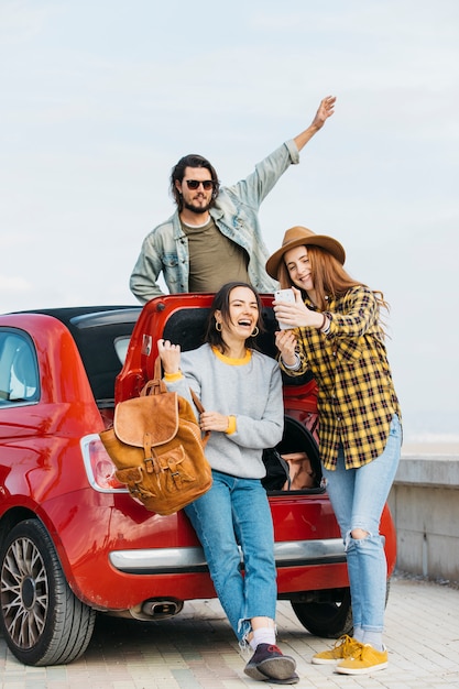 Women taking selfie on smartphone near car trunk and man leaning out from auto