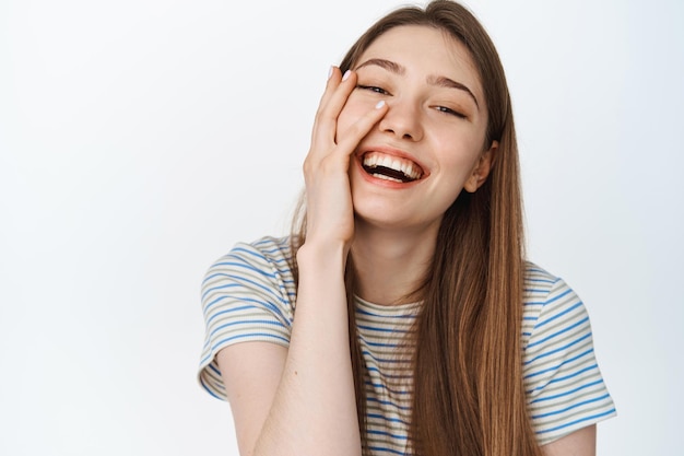 Women wellbeing. Close up of natural beautiful girl, touching clean glowing skin, laughing and smiling, showing white teeth, standing over studio background, isolated.